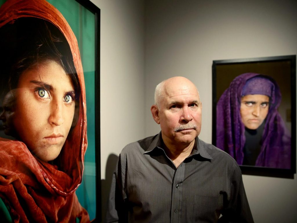 O fotógrafo Steve McCurry entre as duas fotos que fez de Sharbat Gula, uma quando ela era criança e anos depois quando a reencontrou adulta | Foto: Getty